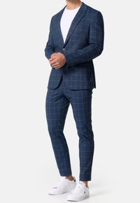 Navy blauw geblokt pak met een blazer met één knoop, slim-fit broeken en witte sneakers. De stof heeft een gladde textuur, met contrasterende ruitenlijnen.