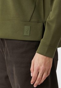 Zbliżenie na rękę osoby odpoczywającą przy boku, ubraną w oliwkową bluzę i ciemno-brązowe spodnie z małą łatką na bluzie.