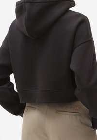 Felpa nera cropped con tasca a marsupio e cappuccio, abbinata a pantaloni beige con una patch con logo sulla tasca posteriore.