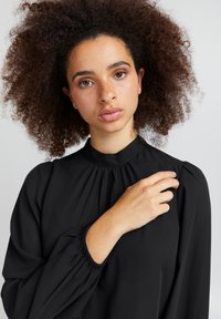Blusa nera realizzata in un tessuto liscio, caratterizzata da un colletto alto, dettagli arricciati e polsini elasticizzati per una vestibilità comoda.