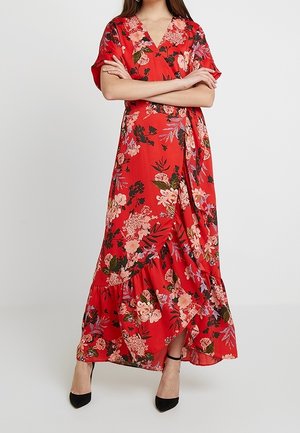 Femme portant une robe portefeuille rouge à imprimé floral avec un ourlet à volants et des talons noirs, les bras croisés, debout devant un fond uni.