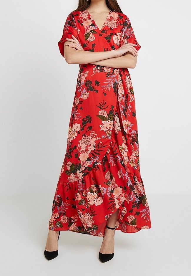 Femme portant une robe portefeuille rouge à imprimé floral avec un ourlet à volants et des talons noirs, les bras croisés, debout devant un fond uni.