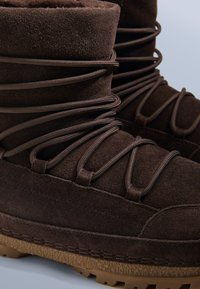 Barna velúr csizma, texturált felülettel, lekerekített teteje és keresztbe fűzött fűzői vannak. Látható gumi talpak tapadási részletekkel.