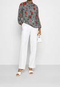 Mujer vestida con una blusa larga de manga larga gris con flores, pantalones anchos blancos, sandalias de tacón blancas, sosteniendo un pequeño bolso blanco.