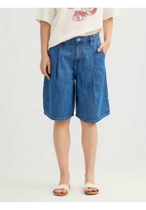 Persona que lleva puestos unos pantalones cortos de mezclilla azul sueltos hasta la rodilla y sandalias trenzadas color crema, con una mano en el bolsillo, sobre un fondo liso.