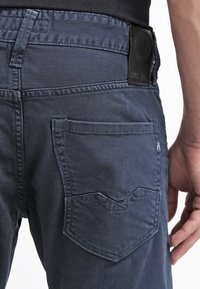 Vue arrière d'une personne portant un jean bleu foncé avec des coutures détaillées sur la poche arrière et un patch de marque en cuir noir sur la ceinture.