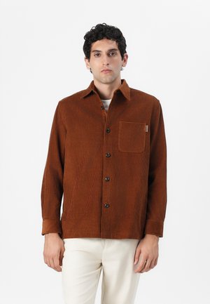 Man wearing a brown corduroy button-up shirt with a front pocket and white pants, standing against a plain white background.