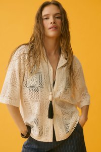 Femme aux longs cheveux mouillés porte une chemise crème en crochet à boutons, un collier à pompons noir et un pantalon sombre à rayures sur fond jaune.