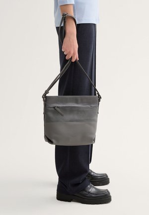Gray leather bucket bag with a textured upper panel, adjustable strap, and a small side pocket, held in one hand.