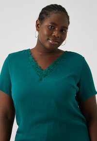 Blouse teal à manches courtes avec un col en V doté d'une bordure en dentelle. Tissu lisse avec un léger éclat et coupe décontractée.