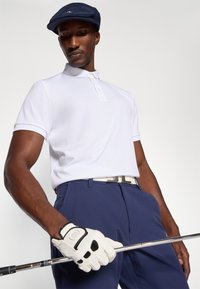 Kortermet hvit polo skjorte, marineblå bukser, hvit belte, og golfhanske. Personen holder en golfkølle og har på seg en marineblå caps.