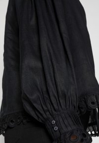 Svart blus med lös passform, samlade ärmar och en dekorativ spetskant med ögonpletter. Knappar pryder ärmslutet. Mjuk tygtextur.