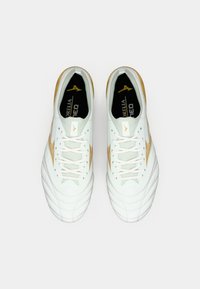 Hvite fotballsko med teksturert overdel, gyldne detaljer og et karakteristisk sikksakkmønster. Har flate hvite lisser og polstret tunge.