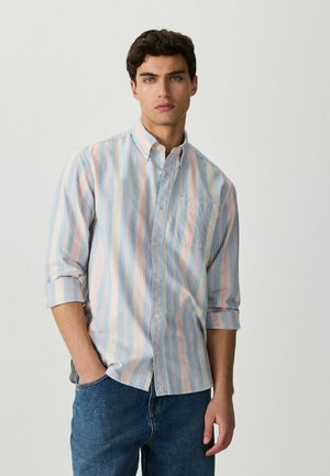 Young man with dark hair wearing a light blue, pink, and white vertically striped button-up shirt and blue jeans, posing with one hand in pocket.