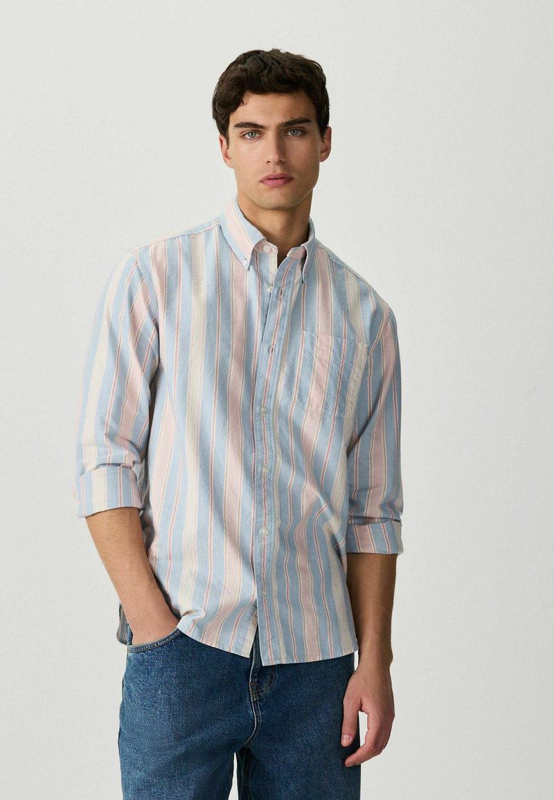 Noor mees tume juustega, kandes helerohelist, roosat ja valget vertikaalselt triibulist nööbitavat särki ja siniseid pükse, poseerides ühe käega taskus.