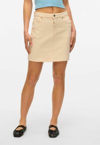 Falda de mezclilla beige con diseño ajustado, bolsillos delanteros y cierre con un solo botón. Se lleva con zapatos planos negros y una blusa de color azul claro.