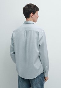 Camicia azzurro chiaro con struttura liscia, maniche lunghe e orlo arrotondato. Presenta un rinforzo posteriore e polsini con un solo bottone.