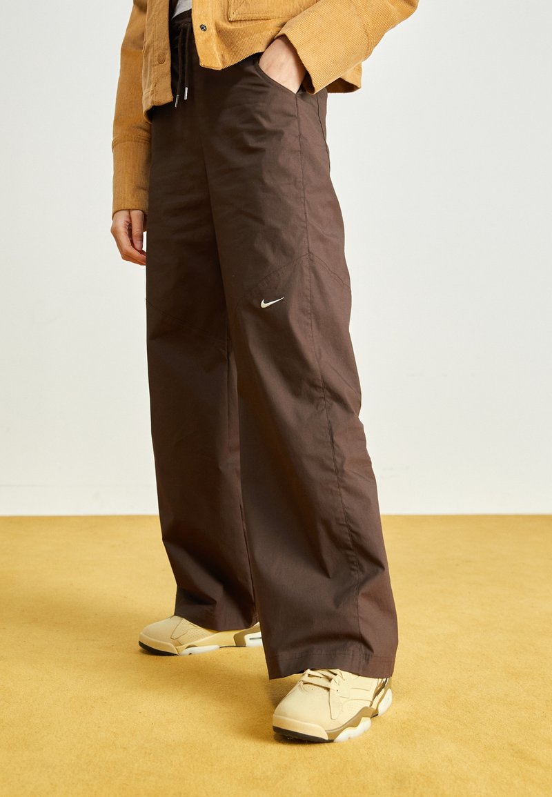 Pantalons brown amples en matériau léger, avec une poche latérale et un petit logo Nike blanc. Portés avec des baskets beiges.