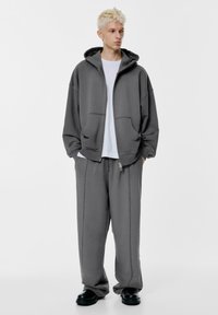 Šedá mikina na zips s veľkou kapucňou, v kombinácii s nohavicami v rovnakom odtieni. Obe súčasti majú viditeľné švy a vrecká. Čierna obuv.