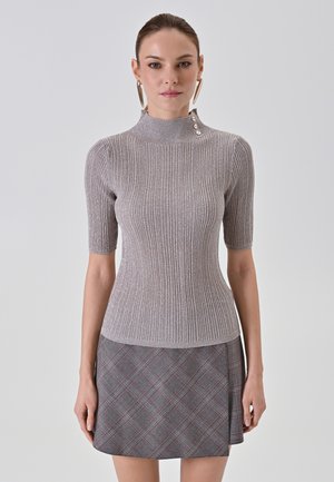 Donna che indossa un top corto a maniche corte aderente in maglia con dettagli a bottoni al collo e una gonna a quadri grigia su uno sfondo neutro.