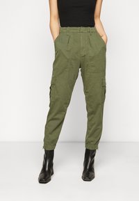 Persoon in olijfgroene cargo broek met hoge taille en zijzakken, met zwarte haklaarzen, staande met de handen in de zakken.