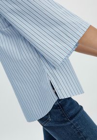Camicia a righe blu chiaro con maniche corte, dotata di spacchi laterali e una vestibilità rilassata. Realizzata in un tessuto morbido con una consistenza liscia.