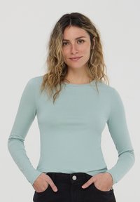 Top fitted a maniche lunghe blu chiaro realizzato in materiale morbido, con un collo rotondo discreto e una texture liscia, abbinato a pantaloni scuri.