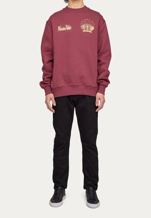 Burgundy sweatshirt med guldbroderi som har en design på vänster bröst och en krona grafisk. Parat med svarta byxor och sneakers.