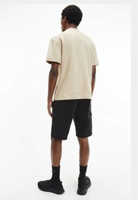 Beżowy t-shirt z okrągłym dekoltem, luźny krój, zestawiony z czarnymi spodniami cargo z bocznymi kieszeniami, noszony z czarnymi butami sportowymi i skarpetkami.