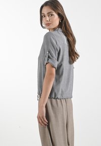Blusa grigia a maniche corte con orlo arricciato, maniche abbottonate e colletto semplice. Tessuto morbido, vestibilità comoda, abbinata a pantaloni marrone chiaro.