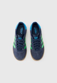 Tengerészkék velúr tornacipő zöld és kék részletekkel, három csíkkal, fűzős elejjel, gumitalppal és párnázott nyelvvel.