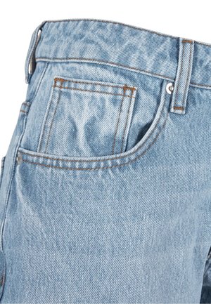 Femme portant un crop top blanc sans manches, un jean bleu clair ample et des baskets grises, debout devant un fond uni.