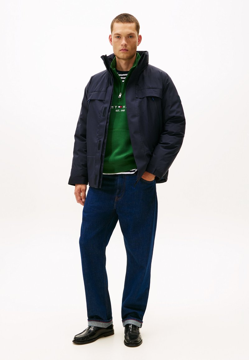 Marinblå puffjacka med hög krage, över en grön hoodie och randig skjorta, kombinerad med blå jeans och svarta skor. Enkel design, funktionell stil.