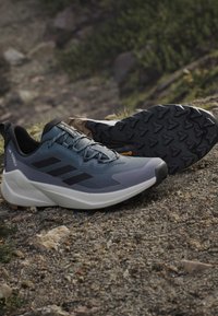 Blaue und graue Sportschuhe mit schwarzen Akzenten, strukturiertem Stoff, einer dicken gepolsterten Sohle und robustem Profil für besseren Halt.