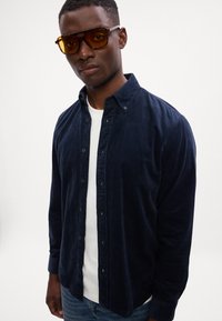 Mørkeblå kordfløjl button-up skjorte over en hvid t-shirt, med en klassisk krave og lange ærmer, parret med oversized gule solbriller.