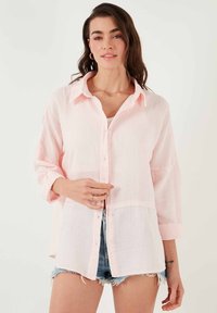 Light pink button-up shirt with a textured fabric. Features a collar, long sleeves rolled to mid-arm, and a relaxed fit. Worn over denim shorts.