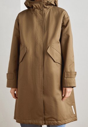 Osoba ubrana w brązowy płaszcz sięgający do kolan z kapturem, kieszeniami z klapami z przodu, zapinanymi paskami na rękawach oraz ukrytym zapięciem z przodu.
