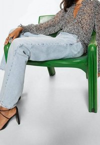 Femme allongée sur une chaise verte, portant un blouse noire à fleurs, un jean bleu clair et des chaussures à talons hauts noires à lanières.