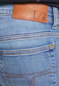 Ljust blå denimjeans med orange sömmar, en brun läderbälte med "T/J", och klassisk femficka design.