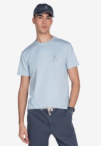 Camiseta de algodón azul claro con mangas cortas, cuello redondo y diseño bordado que presenta una calavera y hachas cruzadas en el pecho.