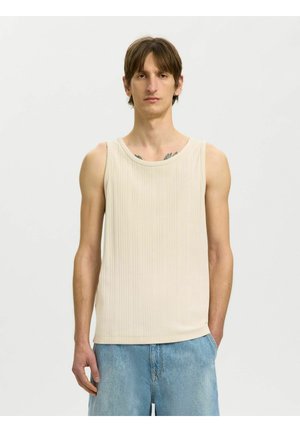 Joven con cabello castaño de longitud media, vistiendo una camisa de canalé beige sin mangas y pantalones cortos de mezclilla azul claro, de pie sobre un fondo liso.