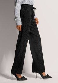 Zwarte, gestructureerde wijde broek met een subtiel camouflagepatroon, voorzien van een elastieken tailleband en trekkoorddetail, gecombineerd met puntige hakken.