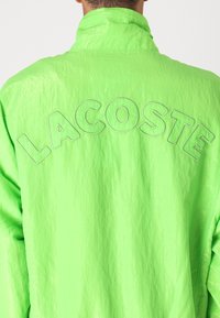 Casaco impermeável verde neon com um tecido texturizado, apresentando um grande logótipo em relevo "LACOSTE" na parte superior das costas. Design de colarinho clássico.