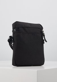 Svart rektangulär crossbody-väska med framficka med dragkedja, tygrem och huvudsakligt fack med dragkedja på vit pedestal.