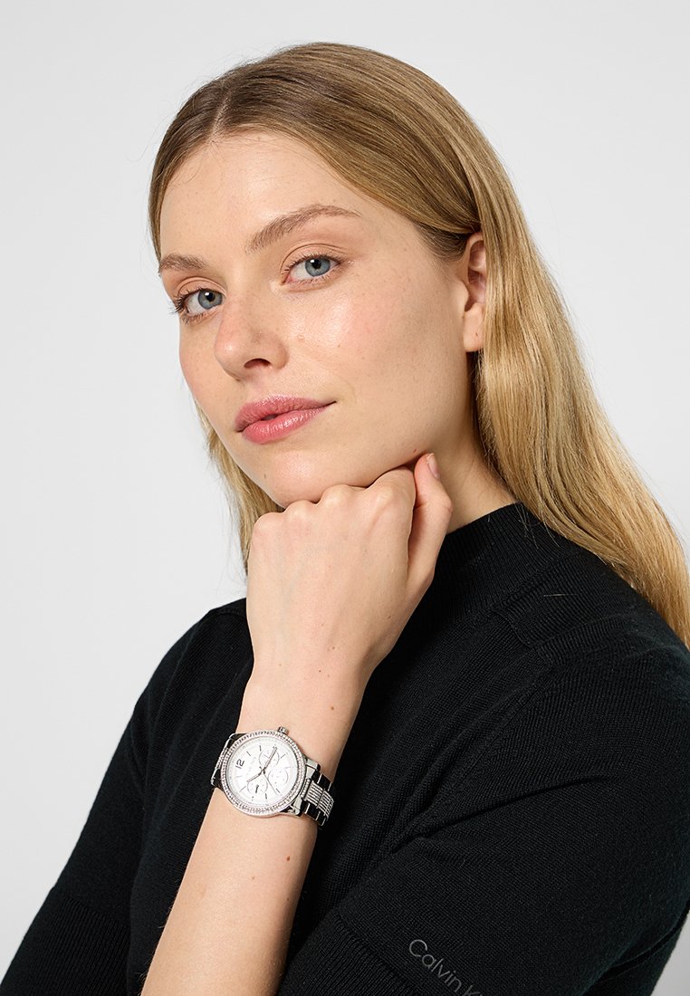 Montre-bracelet en argent avec un cadran rond blanc et des accents en cristal, bracelet en chaîne métallique, présentée sur un poignet avec un haut en tricot noir neutre.