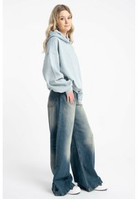 Jasnoniebieska, oversize bluza z kapturem z teksturowym wzorem, zestawiona z szerokimi, przetartymi dżinsami. Ubranie dopełniają białe sneakersy.