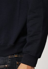 Felpa blu navale con texture morbida, polsini e orlo a coste. Presenta una vestibilità rilassata, cuciture minime ed è abbinata a jeans denim scuri.