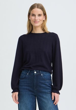 Frau mit blondem Haar, die einen schwarzen, strukturierten Langarmpullover und blaue, hoch taillierte Jeans trägt, steht und lächelt vor einem einfarbigen Hintergrund.