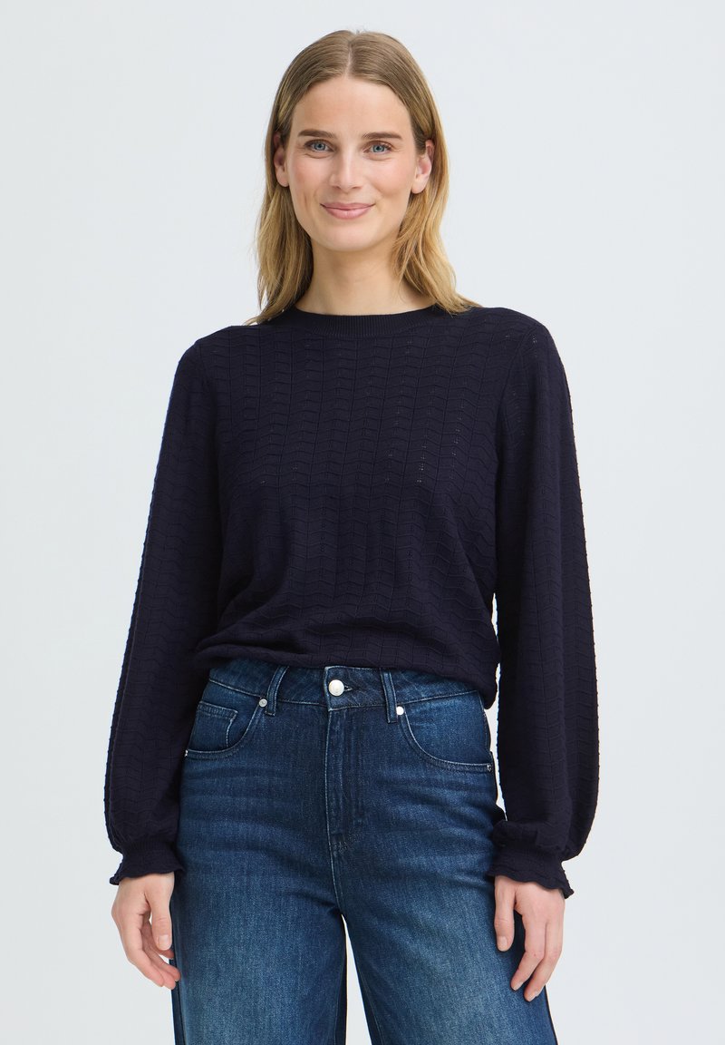Femme aux cheveux blonds portant un pull noir texturé à manches longues et un jean bleu taille haute, debout et souriante sur un fond uni.