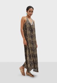 Robe maxi en imprimé léopard avec un décolleté en V profond et des perles élaborées, fabriquée en tissu léger. Le mannequin porte des sandales dorées à lanières.